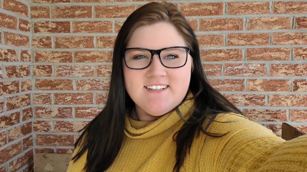 Image of Heather. She is standing infront of brick wall wearing a yellow turtleneck. She has medium length brown hair and square, black rimmed glasses. She is smiling.